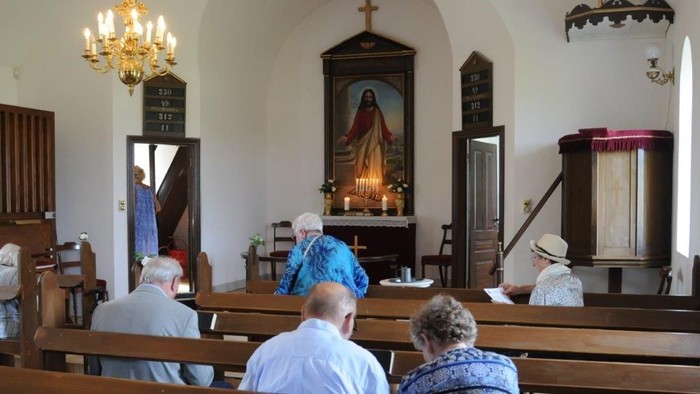 En lille kirke med en liten samling af mennesker, der sidder på bænke og ser mod alteret.