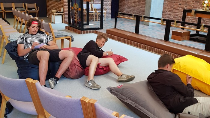 To tre unge mennesker sidder på gulvmatraser i et moderne indretning med mellemliggende stole.