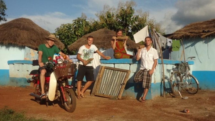Ein Mann sitzt auf einem Motorrad, umgeben von mehreren Personen und Fahrrädern vor einem blauen Haus mit Strohdächern.
