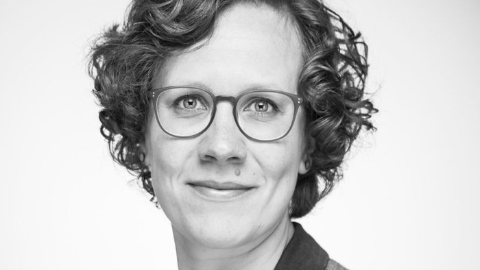 Eine Frau mit kurzem, lockigem Haar und Brille lächelt in die Kamera.