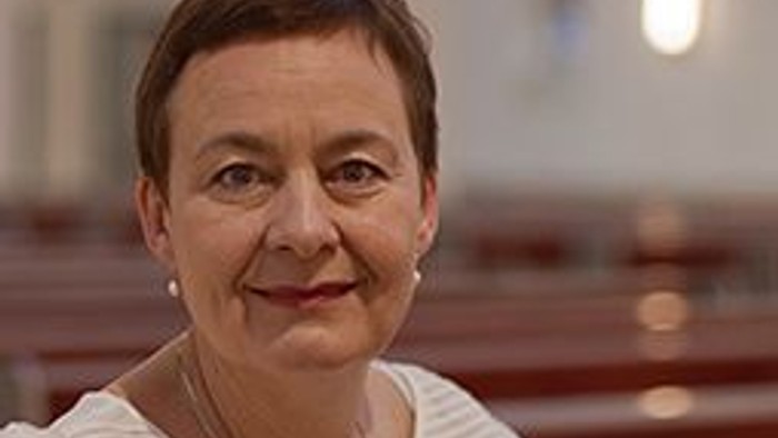 Eine Frau mit kurzen, dunklen Haaren lächelt in einem Raum mit Holzbänken.
