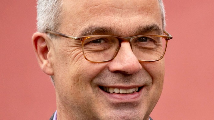 Mann mit Brille und grauen Haaren lächelt vor rotem Hintergrund