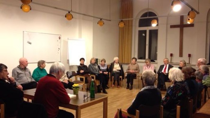 Gruppe von Menschen, die sich in einem gemütlichen Raum mit Holztischen versammelt hat und an einem Meeting oder einer Diskussion teilnimmt.