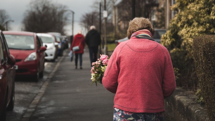 A woman in a pink jacket holds flowers while walking down a sidewalk past parked cars and other pedestrians.