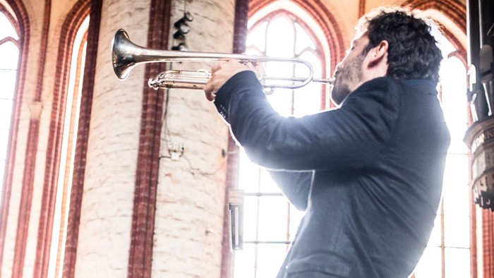 Musiker spielt Trompete, während ein anderer aufmerksam zuhört, in einem prächtigen, sonnendurchfluteten Kircheninneren.