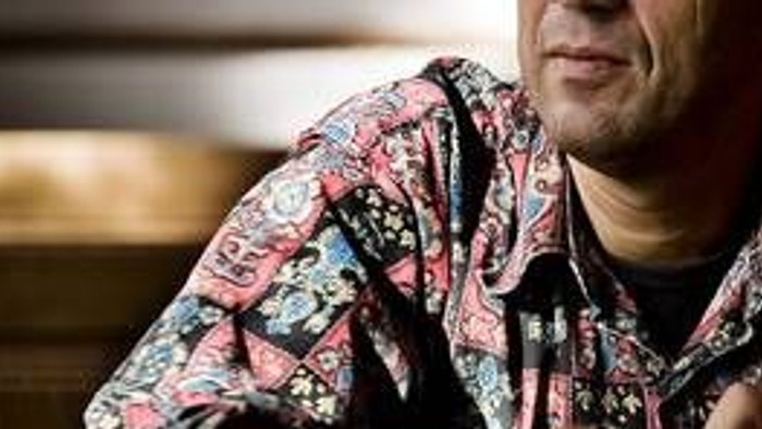 En mand med farverig halsbåndsbluse sidder ved et bord og ser nede i billede.