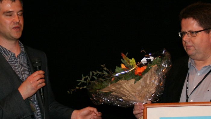 To mænd står på scenen, hvor den ene overrækkes en blomstertop og en præmie.
