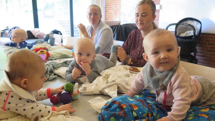 Fire små børn sidder på gulv med tøj og legetøj omkring dem, mens to voksne sitter på en sofa i baggrunden.