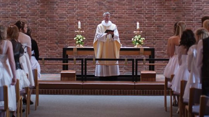 En præst udfører en ceremoniel handling i en moderne kirke med røde mursten og hængende lygter.