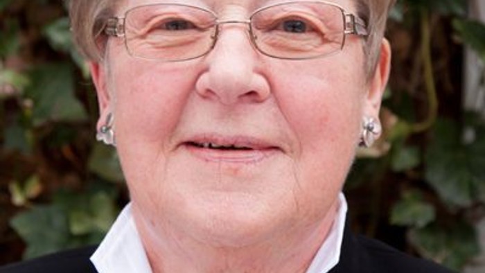 En kvinde med briller og halsbånd står foran et grønt plante.