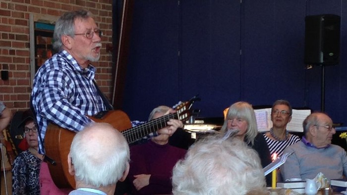 En mand spiller guitar og synger i et lokal, mens andre sidder og ser til.
