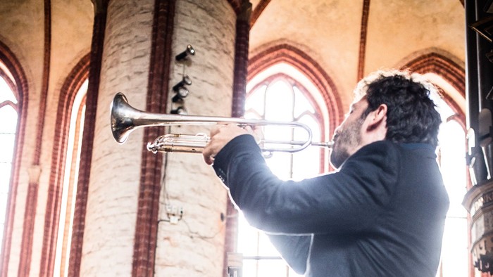 Musiker spielt Trompete, während ein anderer aufmerksam zuhört, in einem prächtigen, sonnendurchfluteten Kircheninneren.
