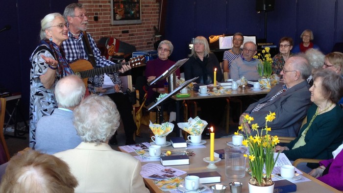 En gruppe ældre folk sidder ved bord med te og blomster.