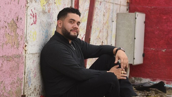 A man with a beard sits against a colorful wall.
