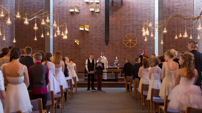 En bryllupsceremoni finder sted i en moderne kirke med lys og gæster i hvidt og rosa.