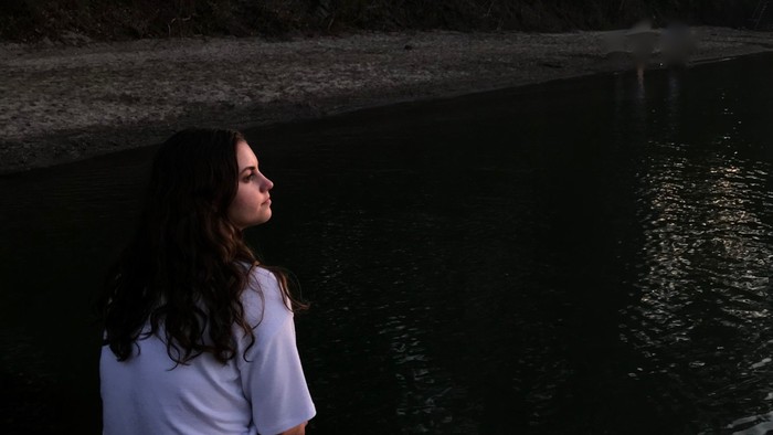 Eine junge Frau sitzt auf einem Felsen am Wasser und schaut nach oben.