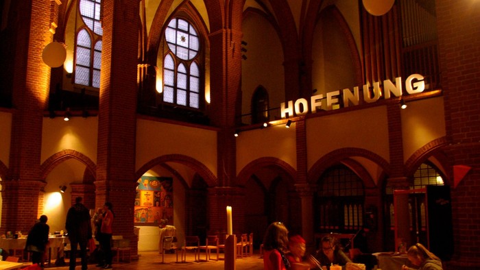 Im Inneren eines großen, gewölbten Raumes sitzen Menschen an Tischen, während ein großes