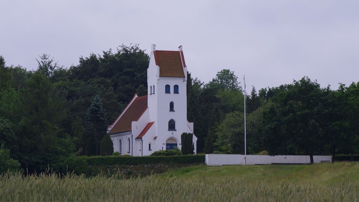 En hvid kirke med røde tag og tårn står i en grøn skov.