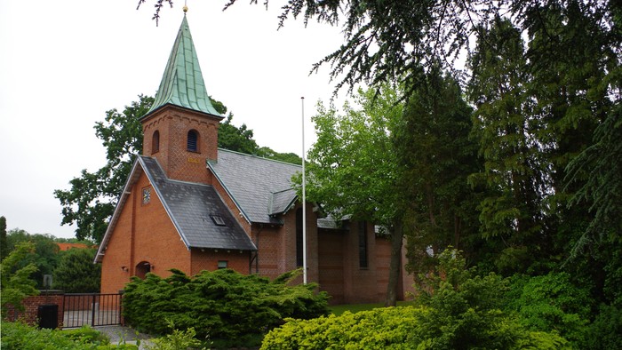 En lille kirke af røde mursten med grøn spire og omgivet af grønne buske og træer