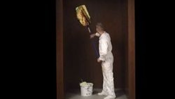 Ein Mann in Overalls hält einen goldenen Rahmen über einen Eimer.