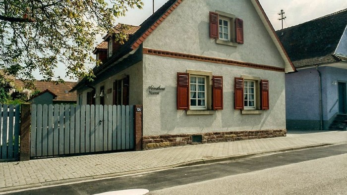 Das Bild zeigt ein charmantes, kleines Haus mit rotem Dach und weißen Fensterläden an einer Kopfsteinpflasterstraße.