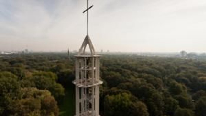 Ein hoher Kirchturm mit Kreuz ragt über einem dichten Wald auf.