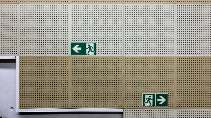 Das Bild zeigt ein Gebäude mit zwei grünen Fluchtwegschildern an einer gelb-beigen Fassade.