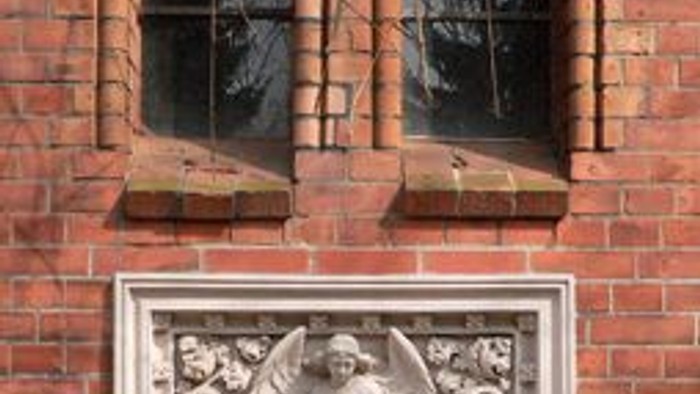 Das Bild zeigt ein steinernes Relief an einer Ziegelmauer unter zwei Fenstern.