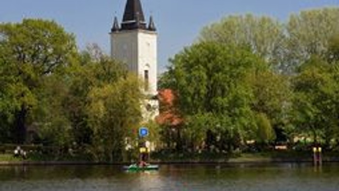 Ein Paar sitzt auf einer Wiese neben Fahrrädern und schaut einem Ruderboot auf einem See zu, im Hintergrund ein Turm.