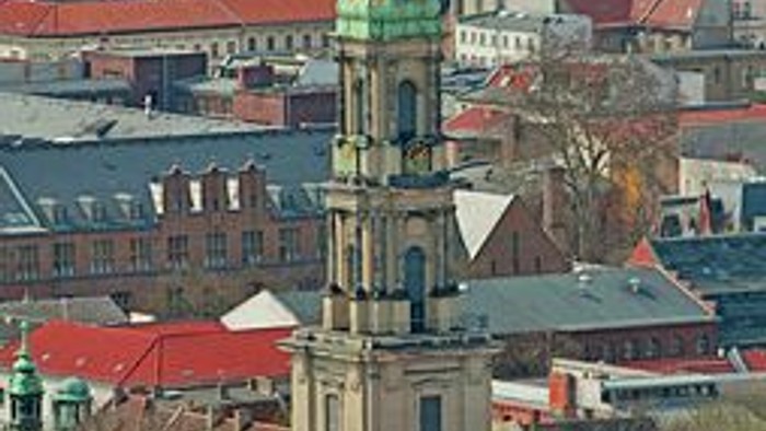 Die Stadtansicht zeigt ein hohes Kirchengebäude mit einem grünen Turm, umgeben von roten und grauen Gebäuden.