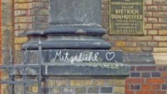 Das Bild zeigt eine Stele mit einer Inschrift und einem Herz, vor einer Ziegelmauer.