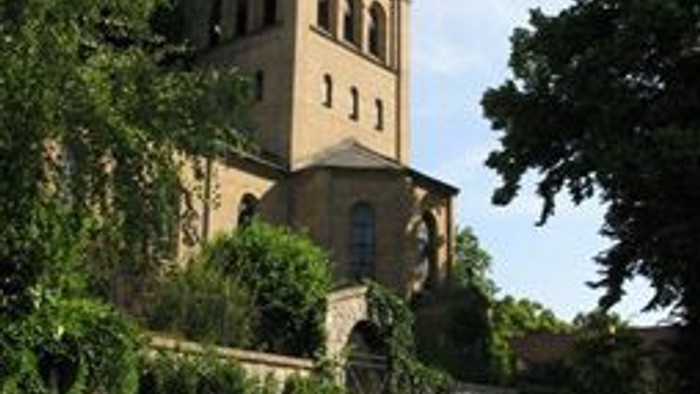 Das Bild zeigt eine alte Kirche mit einem Turm, umgeben von üppigem Grün und einem Kopfsteinpflasterweg.