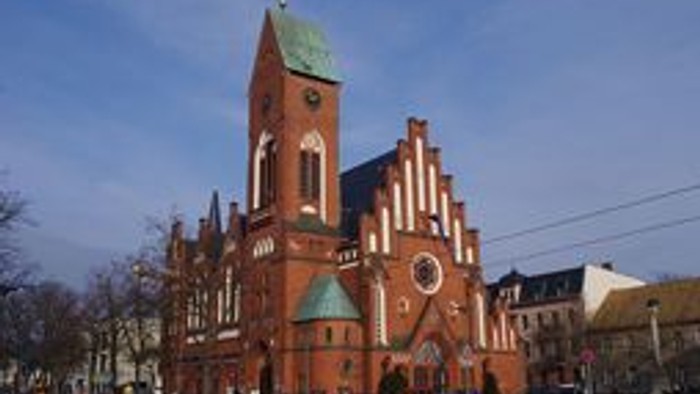 Große rote Kirche mit grünem Dach und mehreren Türmen, vor der Kirche stehen Autos und Menschen.