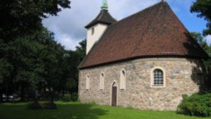 Kleine Kirche mit spitzem Turm und steinernen Wänden, umgeben von grünem Gras und Bäumen