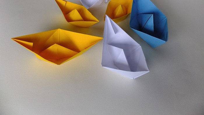 Fünf Origami-Boote in Gelb, Weiß und Blau auf weißem Hintergrund.