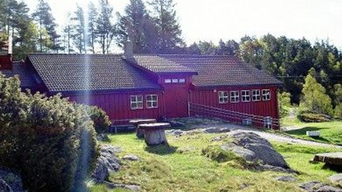 Ein rotes Holzhaus mit Schindeldach liegt in einem grünen, hügeligen Gelände umgeben von Bäumen.