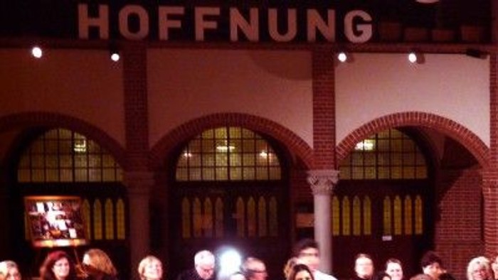 Große Menschenmenge in einem großen Raum mit hohen Bögen und Fenstern, viele stehen und sitzen, einige halten Getränke, ein Fernseher ist an der Wand.