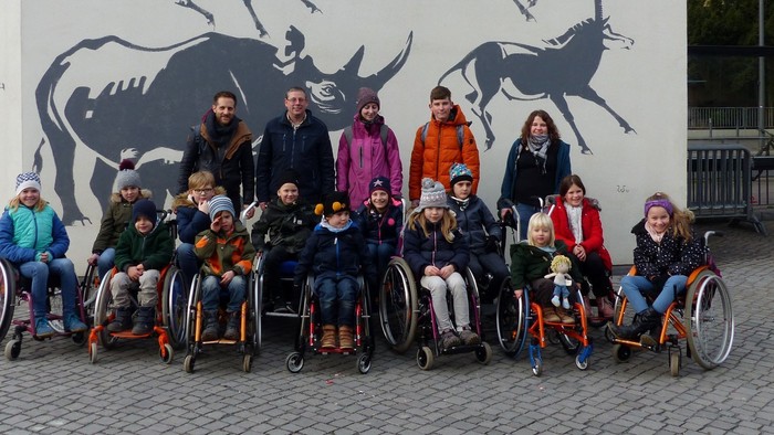 Eine Gruppe von Menschen, einige im Rollstuhl, posiert vor einer Wand mit Kuh-Malerei
