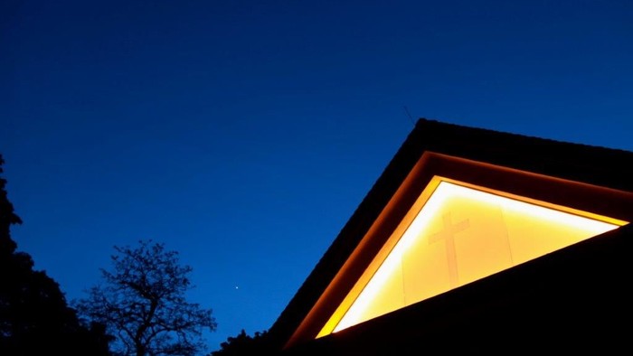 Ein Haus mit beleuchtetem Dachfenster vor dunklem Himmel und Bäumen.