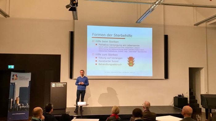 Ein Mann hält einen Vortrag vor einem Publikum, während eine Präsentation hinter ihm auf einem Bildschirm angezeigt wird.