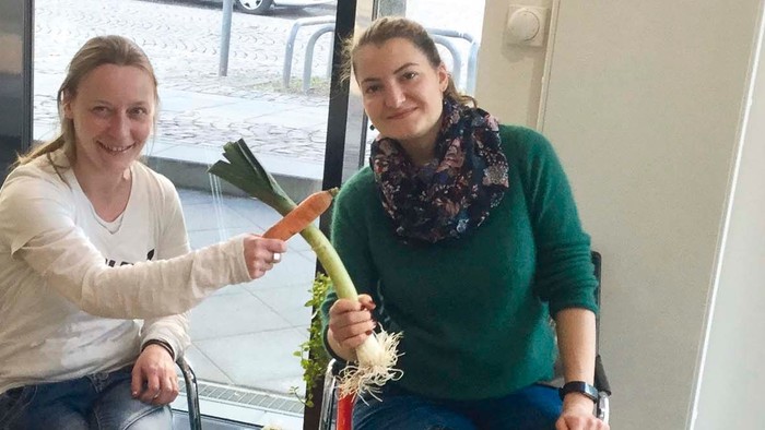 Zwei Frauen sitzen mit Obstkisten und halten Gemüse hoch