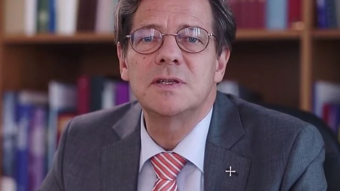 Ein Mann in Anzug und Krawatte sitzt vor Bücherregalen.