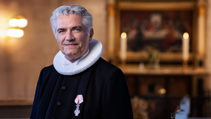 En mand i mørkt tøj og hvid krage står i en rummelig rom med billeder og lysstake på væggen.