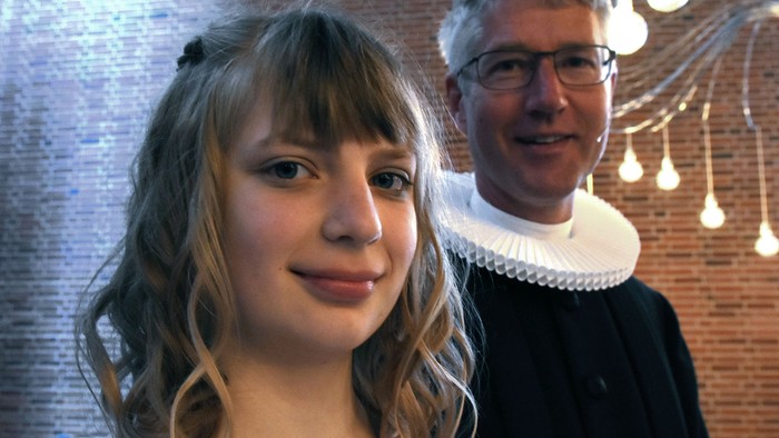En ung pige med blond hår og en mand med briller og hvid krage står sammen.