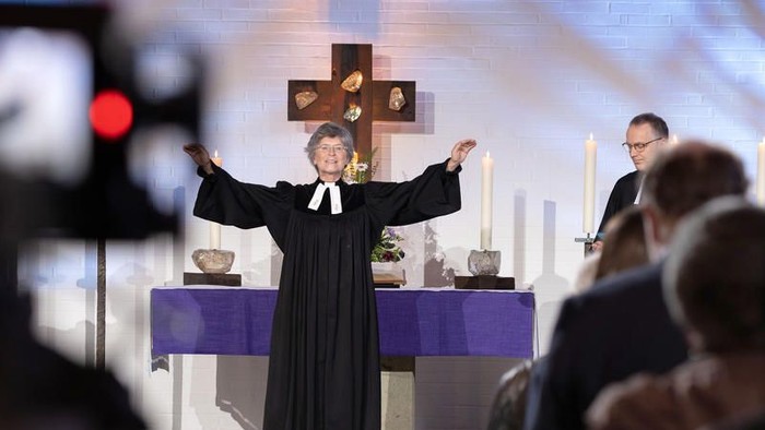 Ein Pfarrer steht mit ausgebreiteten Armen auf der Kanzel während eines Gottesdienstes, umgeben von Gottesdienstbesuchern.