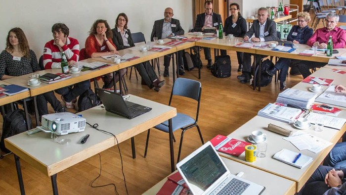 Eine Gruppe von Menschen sitzt an Tischen mit Laptops und Papieren in einem Seminarraum.