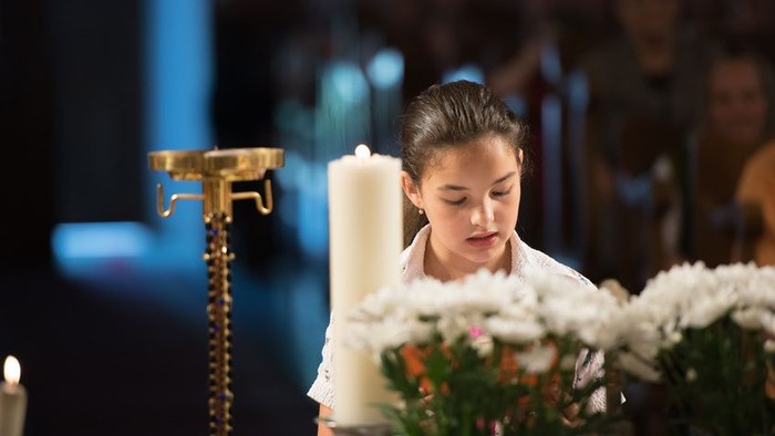 Ein junges Mädchen in weißem Kleid hält eine Kerze und steht vor einem Blumenarrangement.