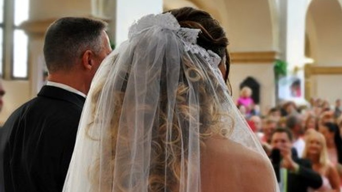 Eine Braut und ihr Vater gehen den Mittelgang einer Kirche entlang, während Gäste zuschauen.