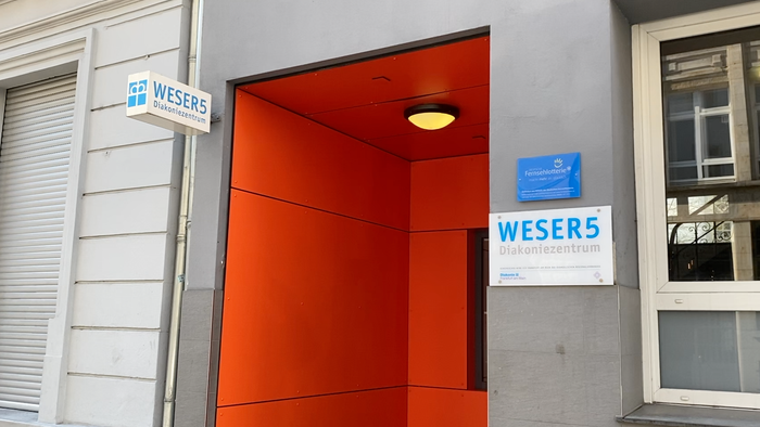 Das Bild zeigt den Eingang eines Gebäudes mit einem orangefarbenen Türrahmen und zwei Schildern mit der Aufschrift