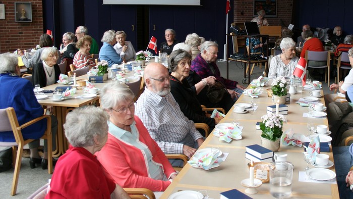 En gruppe ældre samles til et fællesspisning i et lokalet med lange bord og hængelamper.